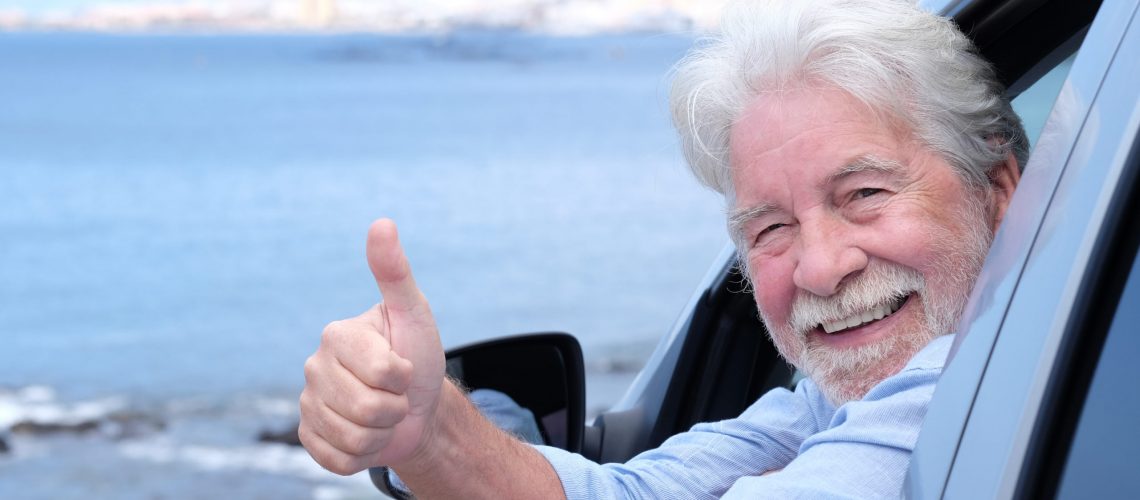 Beautiful white-haired senior man inside the car looking at camera with thumb up. Smiling white haired elderly people enjoying freedom and retirement. Seascape and blue sky